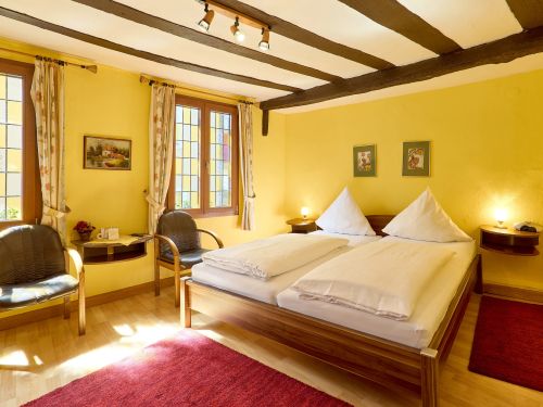 Hell gelbes Schlafzimmer mit Doppelbett zwei Stühlen und Fensterfront mit Gardinen und Holzbalkendecke