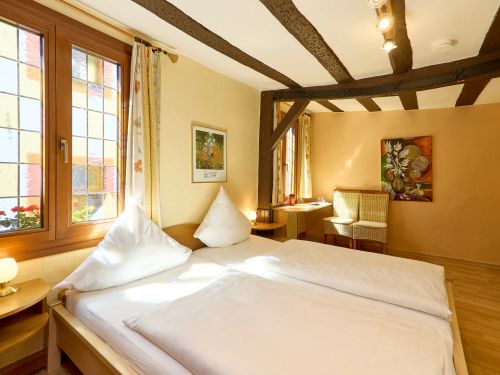 Helles Schlafzimmer mit Doppelbett zwei Stühlen Holzbalken und Fenstern mit Blumen an den Fenstersimsen