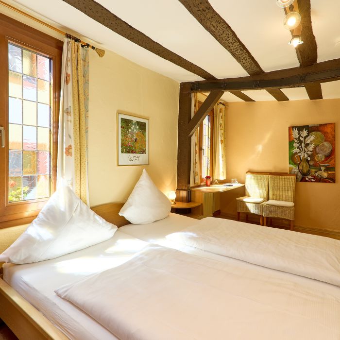 Hell eingerichtetes Schlafzimmer mit großem Bett Holzdeckenbalken und zwei Stühlen am Fenster